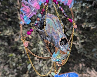 May include: A hanging ornament featuring an iridescent animal skull centered within a gold-toned metal hoop. The skull is adorned with pink and blue aura quartz crystals, colorful beads, and gold chains, with a blue moth cutout suspended below.