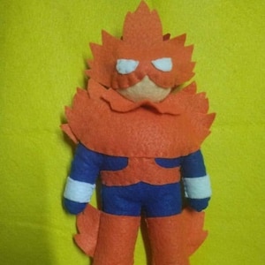 Puede incluir: Un juguete de fieltro hecho a mano de un superhéroe con un traje rojo y naranja y una máscara azul y blanca. El juguete está de pie sobre una superficie amarilla.