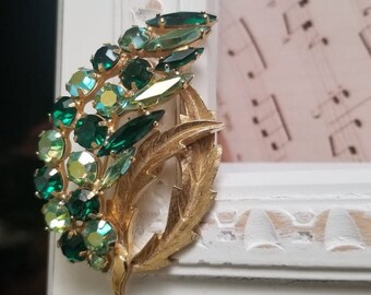 May include: A gold tone brooch with a leaf design and green rhinestones. The brooch is a vintage style piece of jewelry.
