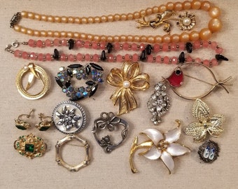 May include: A selection of vintage jewellery including necklaces, brooches, earrings, and a ring. The pieces feature various designs, including pearls, rhinestones, and floral motifs. Colours include gold, pink, green, and silver.