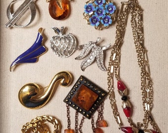 May include: An assortment of vintage brooches and a gold chain necklace. The brooches feature various designs including a pineapple, a cluster of blue flowers, a swirl, a geometric design, and a leaf design. The necklace is made of multiple strands of gold chain with red and black accents.