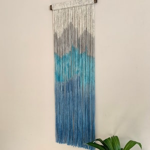 May include: A wall hanging made of yarn with a gradient of white, gray, blue, and teal colors. The yarn is tied to a wooden dowel at the top.