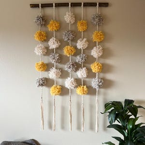 May include: A wall hanging made of yarn pom-poms in shades of yellow, white, and gray. The pom-poms are strung on a natural twine and hang from a wooden dowel. The wall hanging has a rustic and bohemian aesthetic.