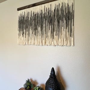 May include: A gray and white yarn wall hanging with a wooden dowel at the top. The hanging is made of a variety of different lengths of yarn, creating a textured and abstract design.