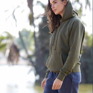 May include: A woman wearing a green hooded sweatshirt and blue denim shorts. She is standing in front of a blurred background of trees and water.