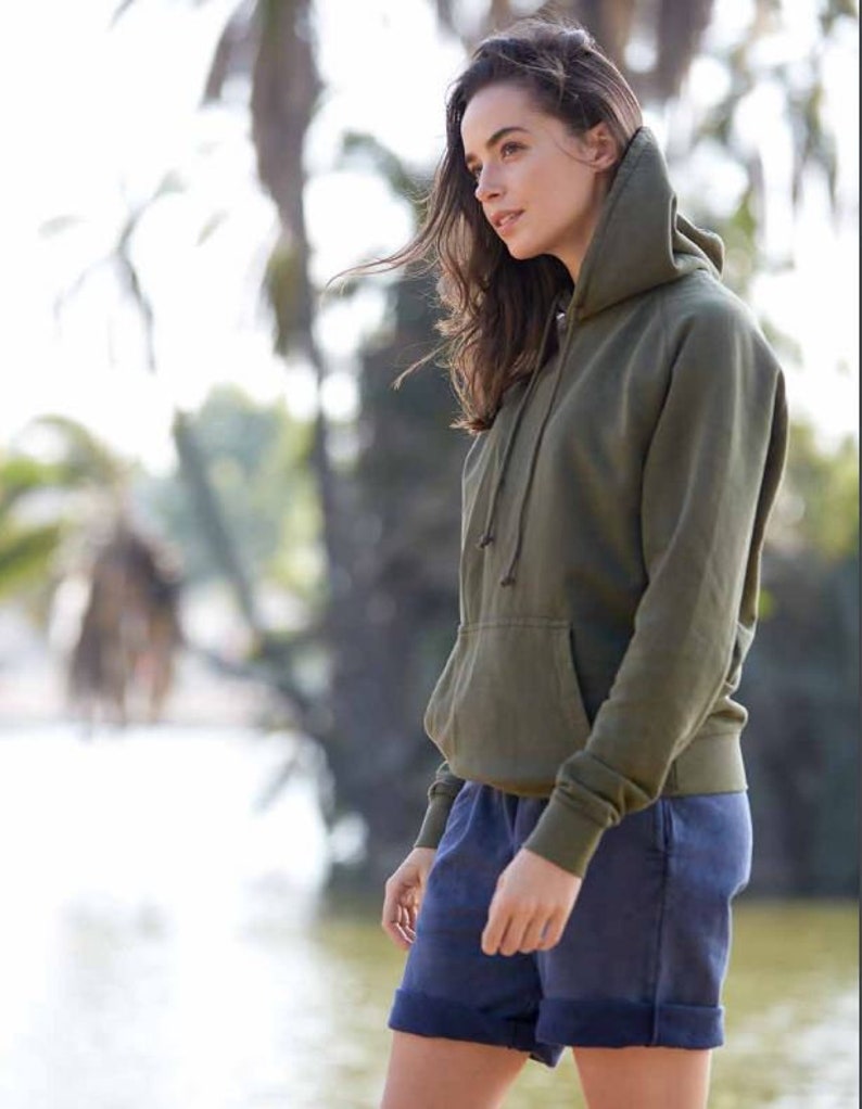 May include: A woman wearing a green hooded sweatshirt and blue denim shorts. She is standing in front of a blurred background of trees and water.