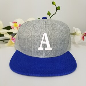 Op de afbeelding: Een grijze en blauwe baseballpet met een witte letter 'A' geborduurd op de voorkant.