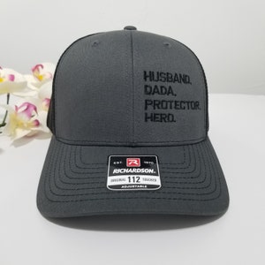 May include: A grey and black trucker hat with the text "HUSBAND. DADA. PROTECTOR. HERO." embroidered on the front. The hat is adjustable and has a Richardson logo on the back.