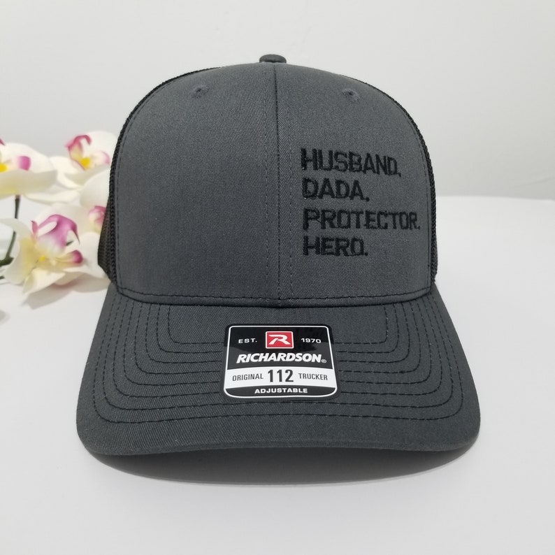 May include: A grey and black trucker hat with the text "HUSBAND. DADA. PROTECTOR. HERO." embroidered on the front. The hat is adjustable and has a Richardson logo on the back.