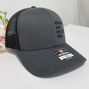 May include: A grey and black trucker hat with the words "Husband, Dada, Protector, Hero" embroidered on the front. The hat is adjustable and has a black mesh back.