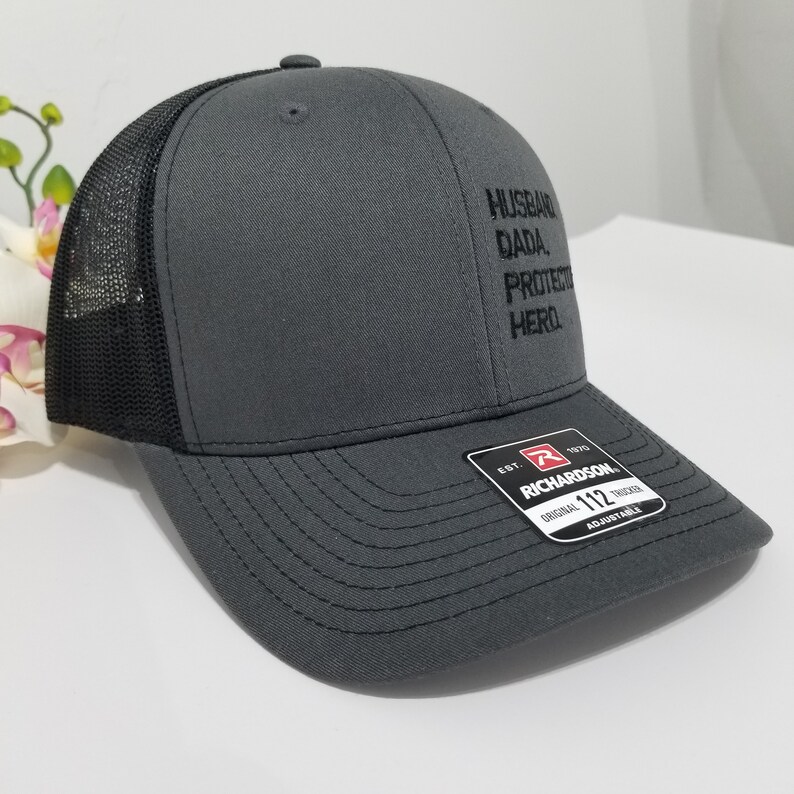 May include: A grey and black trucker hat with the words "Husband, Dada, Protector, Hero" embroidered on the front. The hat is adjustable and has a black mesh back.