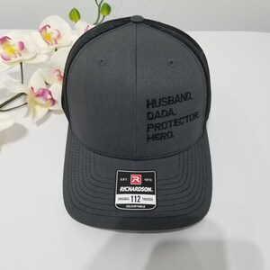 May include: A grey trucker hat with a black mesh back. The hat has black embroidered text that reads "Husband. Dada. Protector. Hero." The hat has a red and black label that reads "Est. 1970 Richardson Original 112 Trucker Adjustable."