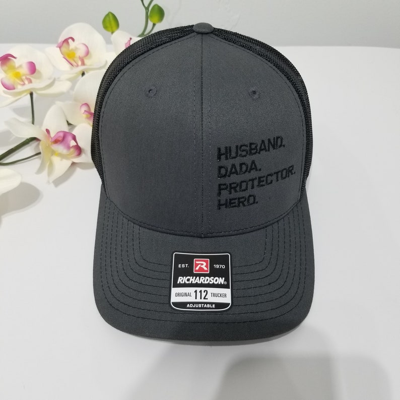 May include: A grey trucker hat with a black mesh back. The hat has black embroidered text that reads "Husband. Dada. Protector. Hero." The hat has a red and black label that reads "Est. 1970 Richardson Original 112 Trucker Adjustable."