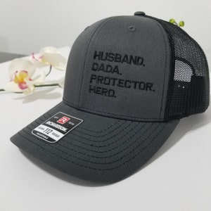 May include: A grey and black trucker hat with the text "Husband, Dada, Protector, Hero." embroidered on the front. The hat is adjustable and has a Richardson 112 label.