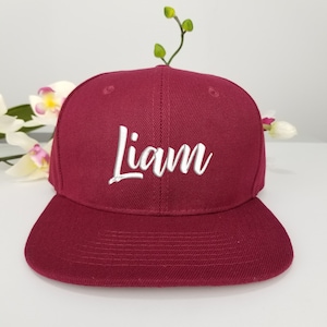 Puede incluir: Una gorra de béisbol de color burdeos con el nombre "Liam" bordado en blanco en la parte delantera.