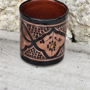 May include: A cylindrical ceramic cup with a brown interior and a decorative exterior. The exterior features a black border at the top and bottom, with a tan background and black floral and dot patterns. The cup is on a concrete surface.