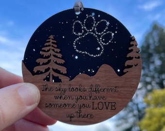 May include: A wooden ornament with a black background and a paw print made of white stars. The ornament has a quote that reads "The sky looks different when you have someone you LOVE up there".