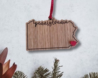 May include: A wooden ornament shaped like the state of Pennsylvania with the word "Pennsylvania" written on it. The ornament has a red heart in the lower right corner and a red ribbon attached to the top.