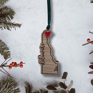 May include: Wooden ornament shaped like the state of Delaware with the word "Delaware" written on it. A red heart is placed on the ornament where Dover, Delaware is located. The ornament is hanging from a green ribbon.