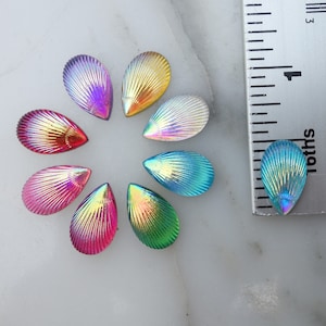 May include: Eight iridescent seashell-shaped glass beads in various colors, including pink, blue, green, and gold. The beads are arranged in a circle on a white surface.