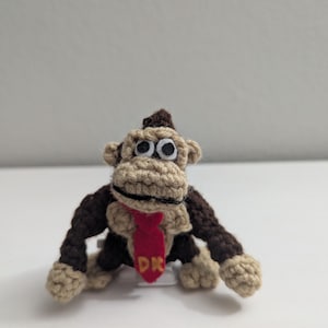 May include: A small crocheted gorilla figure with dark brown and tan yarn. The figure features a red felt necktie with the yellow letters DK embroidered on it, white and black felt eyes, and a wide tan muzzle with a stitched black mouth line.