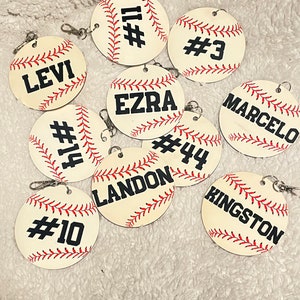 Könnte beinhalten: Zehn Baseball-Schlüsselanhänger mit aufgedruckten Namen und Nummern. Die Schlüsselanhänger sind aus Holz und haben einen weißen Hintergrund mit roten Nähten und schwarzen Buchstaben.