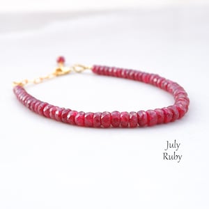 May include: A red ruby gemstone beaded bracelet with a gold clasp. The bracelet is on a white background. July Ruby.