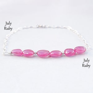 Puede incluir: Un brazalete de cadena de plata con una fila de cinco piedras de rubí ovaladas de color rosa. El brazalete tiene un cierre de mosquetón. El texto "July Ruby" aparece dos veces en la imagen.