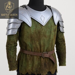 May include: A medieval costume comprising a green tunic and silver armour. The armour includes shoulder pauldrons and a chest plate with a diamond design. A brown belt is fastened at the waist. The costume is displayed on a mannequin.