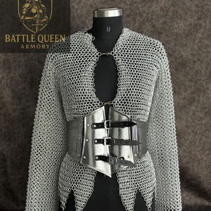 Puede incluir: Una camisa de cota de malla plateada y un cinturón de metal con correas de cuero se exhiben en un maniquí negro. La cota de malla tiene un diseño de cuello en V y está hecha de pequeños anillos de metal entrelazados. El cinturón es de metal y tiene tres hebillas.