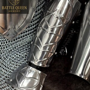 May include: Close-up of polished silver arm armor with leaf-like designs. The armor is part of a set, with chainmail visible in the background. The Battle Queen Armory logo is in the upper left corner.