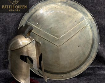 Puede incluir: Un conjunto de casco y escudo espartano de color bronce. El casco tiene una frente y protectores de mejillas distintivos. El escudo es redondo con un diseño en forma de V. El logotipo de Battle Queen Armory está en la esquina superior izquierda.