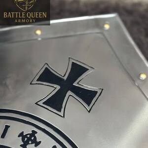 Puede incluir: Primer plano de una placa de metal plateado con un dise&ntilde;o de cruz negra y un emblema circular. La placa tiene un acabado de metal cepillado y peque&ntilde;os remaches dorados. El logotipo "BATTLE QUEEN ARMORY" es visible en la esquina superior izquierda.