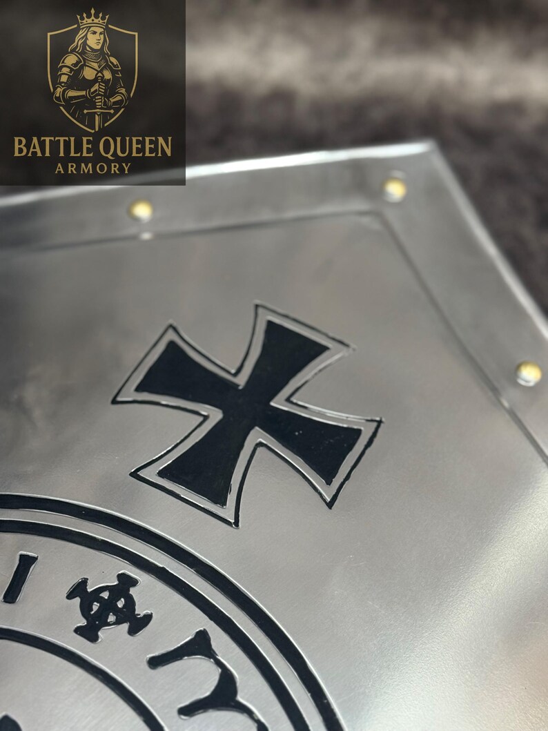 Puede incluir: Primer plano de una placa de metal plateado con un dise&ntilde;o de cruz negra y un emblema circular. La placa tiene un acabado de metal cepillado y peque&ntilde;os remaches dorados. El logotipo "BATTLE QUEEN ARMORY" es visible en la esquina superior izquierda.