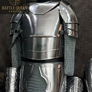 May include: A full suit of polished steel armor, including a breastplate, shoulder guards, and leg protection. Chainmail sleeves and lower body protection are also visible. The armor is displayed on a mannequin, showcasing its medieval design.