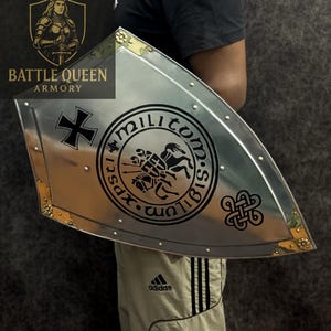 Puede incluir: Un escudo plateado con una cruz negra y un emblema circular con un caballero a caballo. El escudo tiene detalles dorados en las esquinas y est&aacute; remachado. El texto "Battle Queen Armory" es visible.
