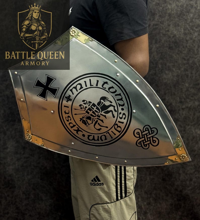 Puede incluir: Un escudo plateado con una cruz negra y un emblema circular con un caballero a caballo. El escudo tiene detalles dorados en las esquinas y est&aacute; remachado. El texto "Battle Queen Armory" es visible.
