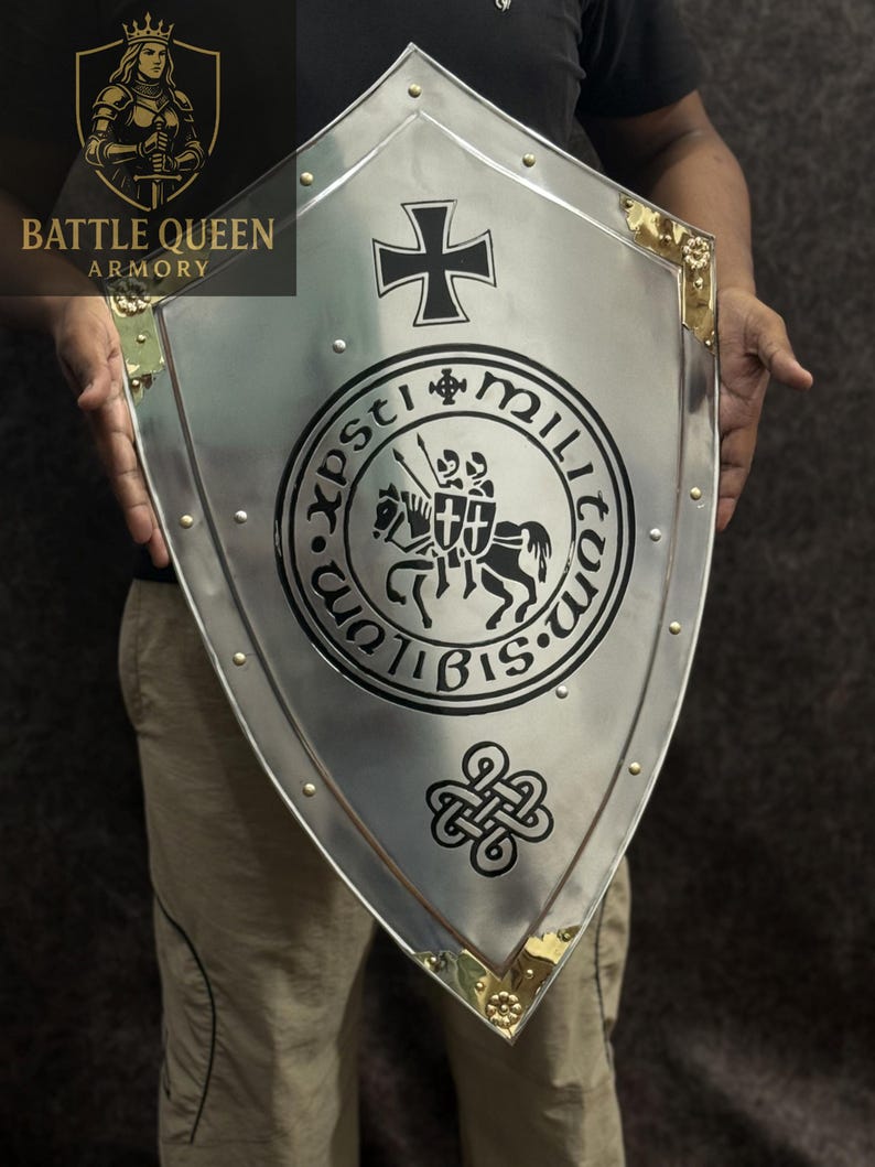 Puede incluir: Un escudo plateado con una cruz negra y un emblema circular con dos caballeros a caballo. El escudo tiene detalles dorados en las esquinas y est&aacute; remachado en los bordes. Se ven las palabras "Battle Queen Armory". Objeto hist&oacute;rico.