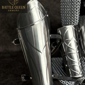 May include: Polished steel arm guards with decorative etched lines and leather straps. The arm guards are part of a set of armor. The Battle Queen Armory logo is visible in the upper left corner.