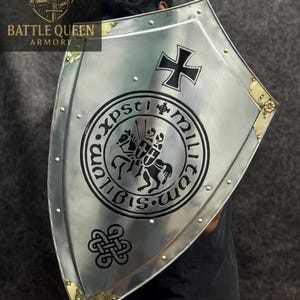 Puede incluir: Un escudo plateado con una cruz negra y un emblema circular con dos caballeros a caballo. El escudo tiene detalles dorados en las esquinas y est&aacute; remachado en los bordes. Se ven las palabras "BATTLE QUEEN ARMORY".