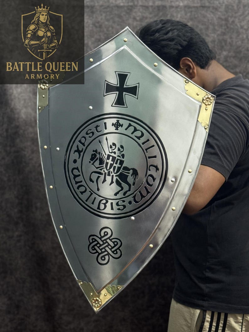 Puede incluir: Un escudo plateado con una cruz negra y un emblema circular. El escudo tiene detalles dorados en las esquinas y remaches. Las palabras "BATTLE QUEEN ARMORY" son visibles en la esquina superior izquierda.
