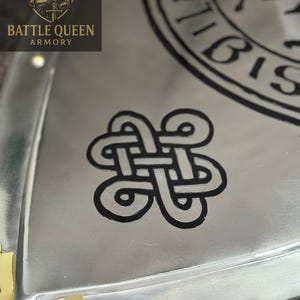 Puede incluir: Primer plano de un objeto met&aacute;lico plateado con un dise&ntilde;o de nudo celta negro. El objeto tiene un acabado pulido y detalles dorados en las esquinas. El logotipo de Battle Queen Armory es visible en la esquina superior izquierda.