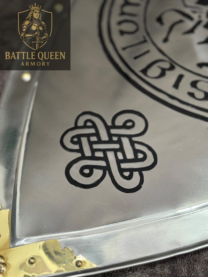 Puede incluir: Primer plano de un objeto met&aacute;lico plateado con un dise&ntilde;o de nudo celta negro. El objeto tiene un acabado pulido y detalles dorados en las esquinas. El logotipo de Battle Queen Armory es visible en la esquina superior izquierda.