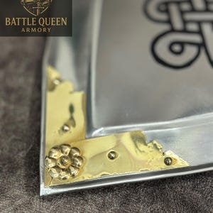 Puede incluir: Primer plano de una bandeja met&aacute;lica con detalles de esquina de lat&oacute;n y un adorno floral. La bandeja tiene un acabado plateado y un dise&ntilde;o de nudo celta. El logotipo "BATTLE QUEEN ARMORY" es visible en la esquina superior izquierda.