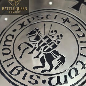 Puede incluir: Primer plano de un escudo de metal plateado con un emblema negro que representa a un caballero a caballo y el texto "Xpsti". El escudo tambi&eacute;n tiene las palabras "Battle Queen Armory" y un logotipo de una reina.