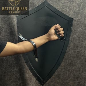 Puede incluir: Escudo negro con la parte superior curvada y la inferior puntiaguda, sujeto por una correa de cuero negro. El escudo tiene un borde remachado. El logotipo de Battle Queen Armory est&aacute; en la esquina superior izquierda.
