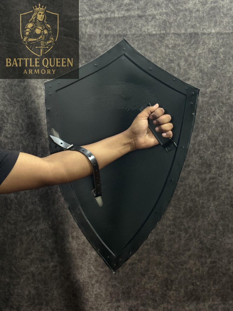 Puede incluir: Escudo negro con la parte superior curvada y la inferior puntiaguda, sujeto por una correa de cuero negro. El escudo tiene un borde remachado. El logotipo de Battle Queen Armory est&aacute; en la esquina superior izquierda.