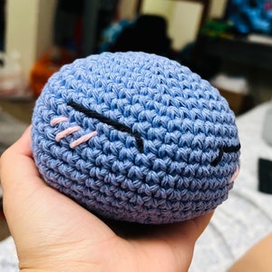 Op de afbeelding: Een blauwe gehaakte bal met geborduurde ogen. De ogen zijn zwart met roze wimpers.