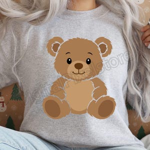May include: A light gray sweatshirt featuring a cartoon teddy bear. The bear is brown with a lighter brown belly and paws, and has a smiling face. The sweatshirt is a casual style.