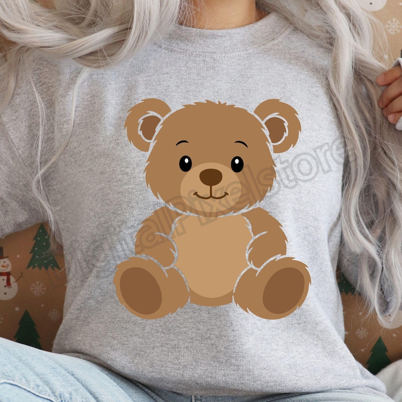 May include: A light gray sweatshirt featuring a cartoon teddy bear. The bear is brown with a lighter brown belly and paws, and has a smiling face. The sweatshirt is a casual style.